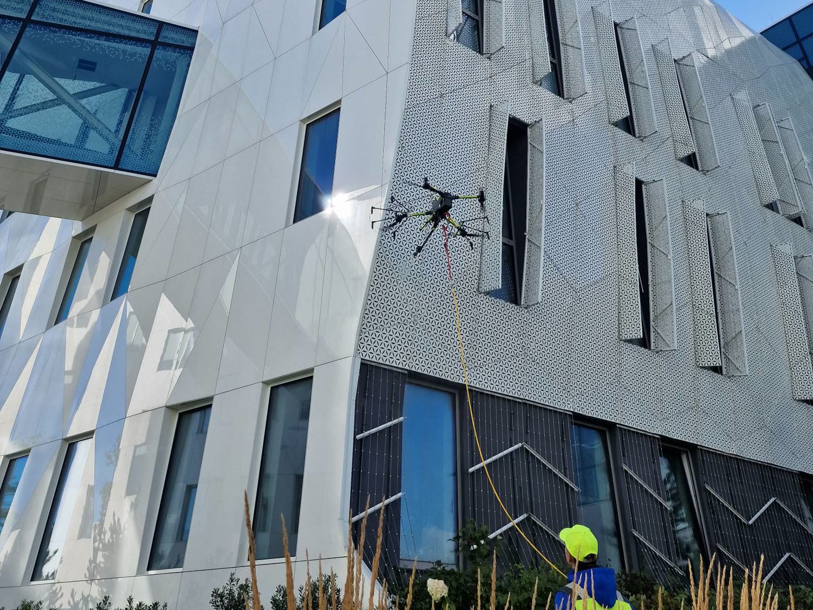 Dron myjący fasadę budynku z przeszkleniami, operator na ziemi